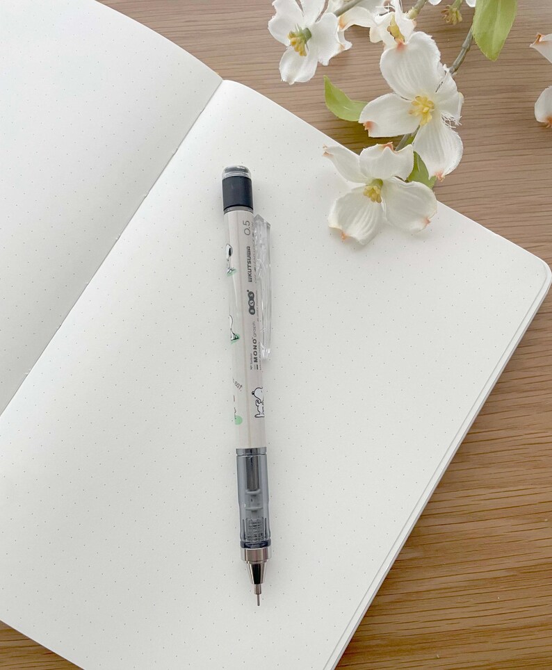 以下が含まれることがあります： 白いノートの上に銀色のシャープペンシルが置かれている。ノートは点線のグリッドパターンで、ペンシルは緑と白のデザインで、側面に「uni-ball signo 0.5」と印刷されている。