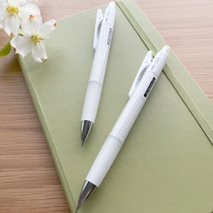 May include: Two white mechanical pencils with a silver clip and a green notebook. The pencils have the text "PENCIL OPL 1.0" printed on the side.