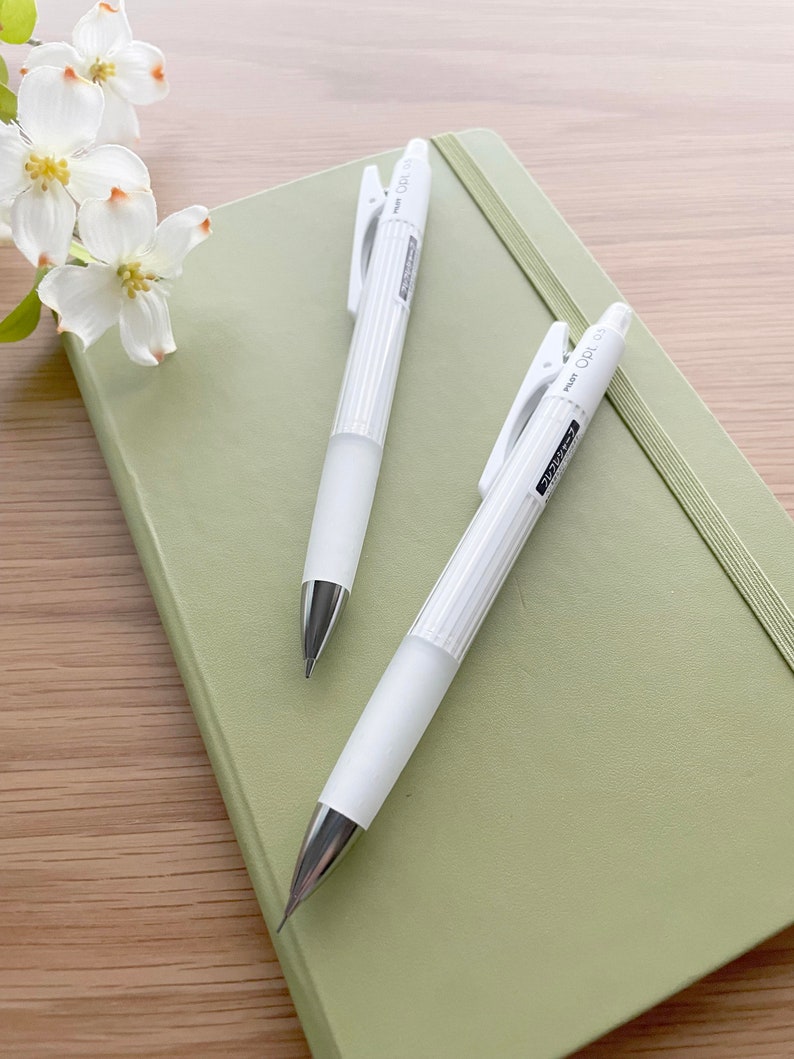 May include: Two white mechanical pencils with a silver clip and a green notebook. The pencils have the text "PENCIL OPL 1.0" printed on the side.