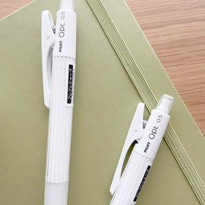 May include: Two white mechanical pencils with a clip on the top. The pencils are labeled "PILOT Opt. 0.5" and have Japanese text on the side. The pencils are on top of a green notebook.