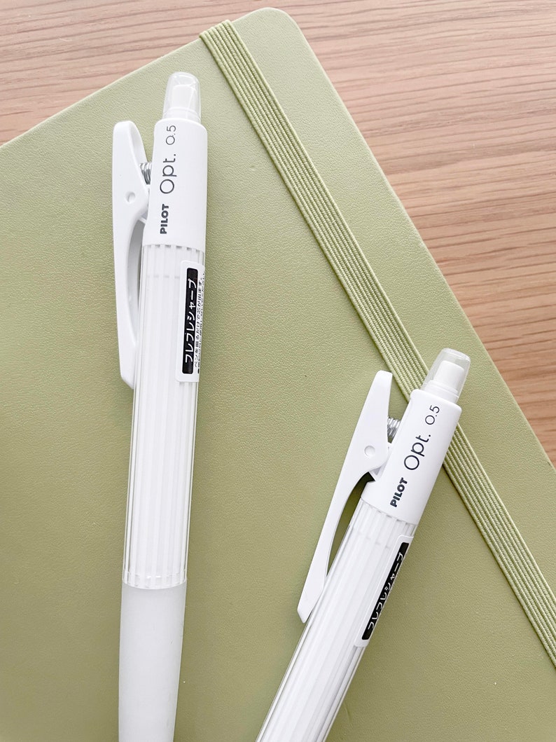 May include: Two white mechanical pencils with a clip on the top. The pencils are labeled "PILOT Opt. 0.5" and have Japanese text on the side. The pencils are on top of a green notebook.