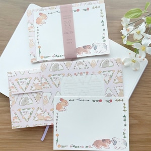 May include: A set of stationery featuring a pink envelope with a repeating pattern of bunnies and green leaves. The set also includes two white cards with bunny illustrations and floral borders. The text "Schnute & Whiskers" and "Letter Set" are printed on the cards.