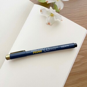May include: A blue and gold Sakura Micron Pigma 03 pen on a white gridded notebook. The pen is labeled "Sakura Micron Pigma 03" and "Micro pigment ink for waterproof and fade proof fine lines". The pen also has the text "水性耐水性" in Japanese.
