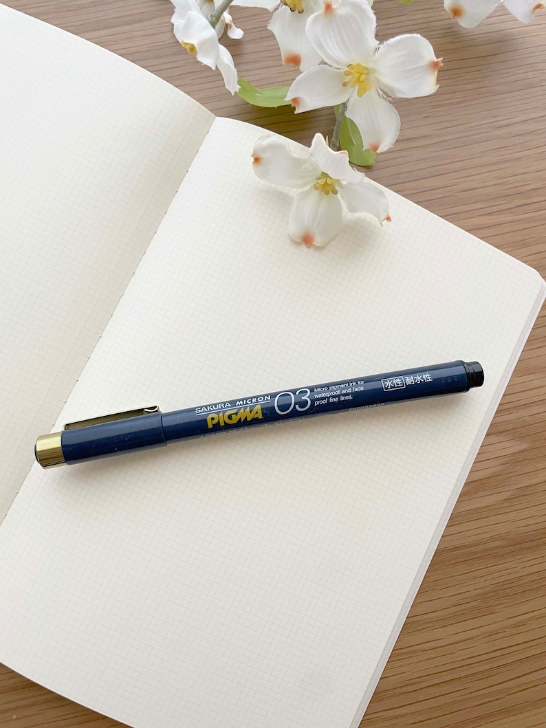 May include: A blue and gold Sakura Micron Pigma 03 pen on a white gridded notebook. The pen is labeled "Sakura Micron Pigma 03" and "Micro pigment ink for waterproof and fade proof fine lines". The pen also has the text "水性耐水性" in Japanese.