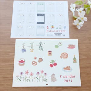 May include: A 2027 wall calendar featuring two sheets with illustrations. The top sheet displays twelve monthly grids with small icons. The bottom sheet shows various watercolor-style drawings of flowers, herbs, food, and a rabbit, with the text Calendar 2027.