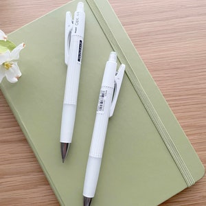 May include: Two white mechanical pencils with a green notebook. The pencils have a silver clip and a white barrel with black text that reads "PENCIL 0.5".