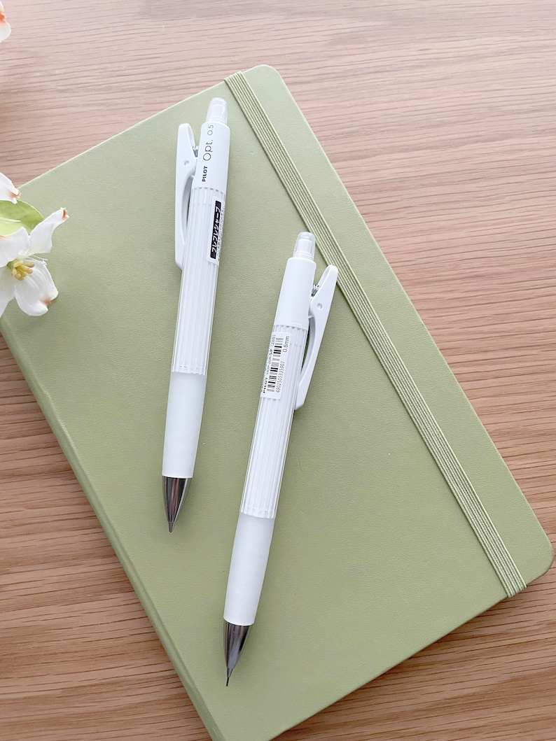 May include: Two white mechanical pencils with a green notebook. The pencils have a silver clip and a white barrel with black text that reads "PENCIL 0.5".