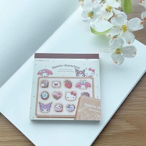 May include: A notepad with a brown background featuring Sanrio characters and chocolates. Each box of yummy chocolates fills the heart with happy. Sanrio characters chocolates.
