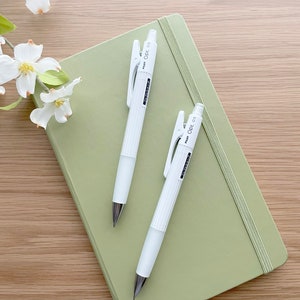 May include: Two white mechanical pencils with a green clip and the text "PENCIL 0.5" on the side, lying on top of a sage green notebook.