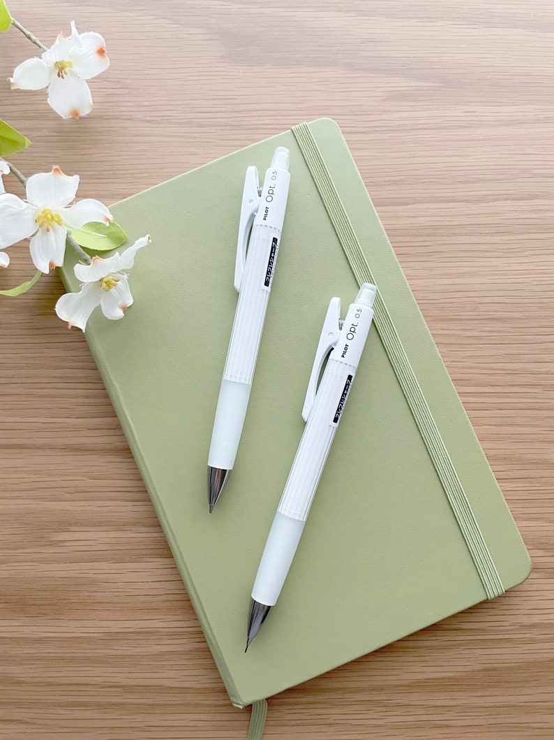 May include: Two white mechanical pencils with a green clip and the text "PENCIL 0.5" on the side, lying on top of a sage green notebook.