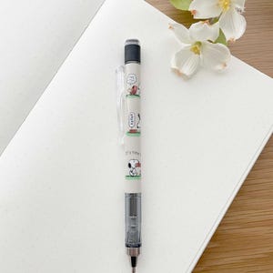 以下が含まれることがあります： 透明なプラスチック製のグリップと黒い消しゴムが付いた白い機械式鉛筆。鉛筆には、ピーナッツのキャラクターであるスヌーピーの漫画のイラストが描かれており、バレルには「It's time to play」という文字が書かれています。