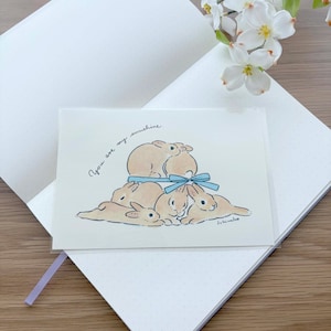 May include: A greeting card featuring a hand-drawn illustration of several tan bunnies with a blue bow. The card is on a white notebook, with the text "You are my sunshine".