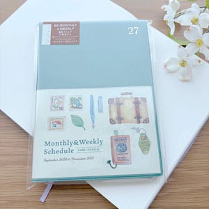 May include: A B6 monthly and weekly schedule journal with a sage green cover. The front features illustrations of postage stamps, a fountain pen, a suitcase, a notebook with a cat graphic, and a key tag. Text reads Monthly & Weekly Schedule.