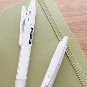 May include: Two white Pilot Opt. 0.5 mechanical pencils with a green notebook in the background. The pencils have a ribbed design and a clip on the top.