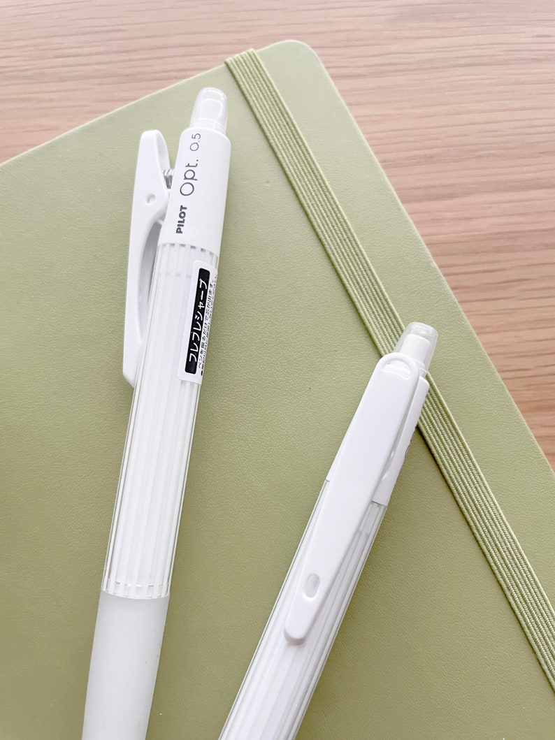 May include: Two white Pilot Opt. 0.5 mechanical pencils with a green notebook in the background. The pencils have a ribbed design and a clip on the top.