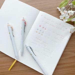 May include: Three white pens with colourful accents and a notebook with handwritten notes about the pens. The notes mention vibrant colours, smooth writing, and a gold tone.