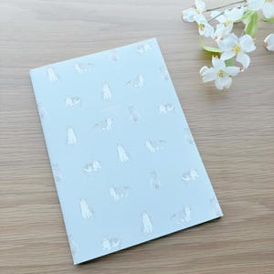 May include: A light blue notebook with a pattern of various cat illustrations. The word "notebook" is printed in the center. The notebook is on a wooden surface, with white flowers in the upper right corner.
