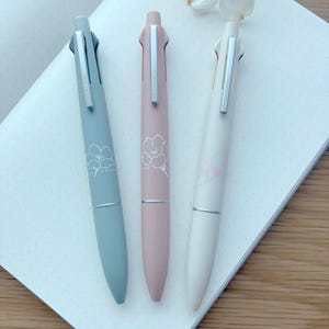 Peut inclure: Trois stylos multicolores aux teintes pastel de bleu, rose et crème, chacun avec un clip argenté et un motif floral. Les stylos reposent sur un cahier blanc à pois, avec une fleur en arrière-plan.