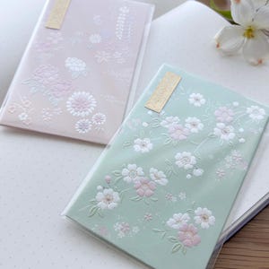 May include: Two decorative Japanese-style notebooks with floral designs. One notebook is pale pink with white and light pink flowers. The other is mint green with white and pink blossoms. Both have a small gold label.