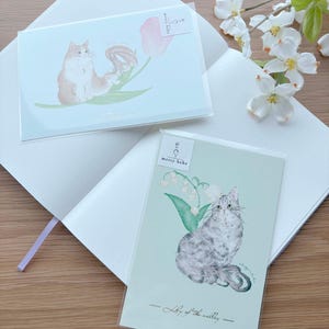 May include: Two greeting cards featuring cat illustrations. One card shows a fluffy orange and white cat with a pink tulip. The other card depicts a gray and white cat with lily of the valley. Both cards have the text "merry bebe".