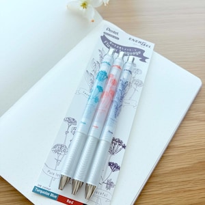 May include: Three silver Pentel EnerGel pens with floral designs and the text "Flower language" on a white card. The pens have turquoise blue, red, and blue black ink. The pens are on a white dotted notebook.