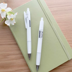 May include: Two white mechanical pencils with a green notebook. The pencils have a black clip and a black inscription that reads "Pilot Opt. 0.5".