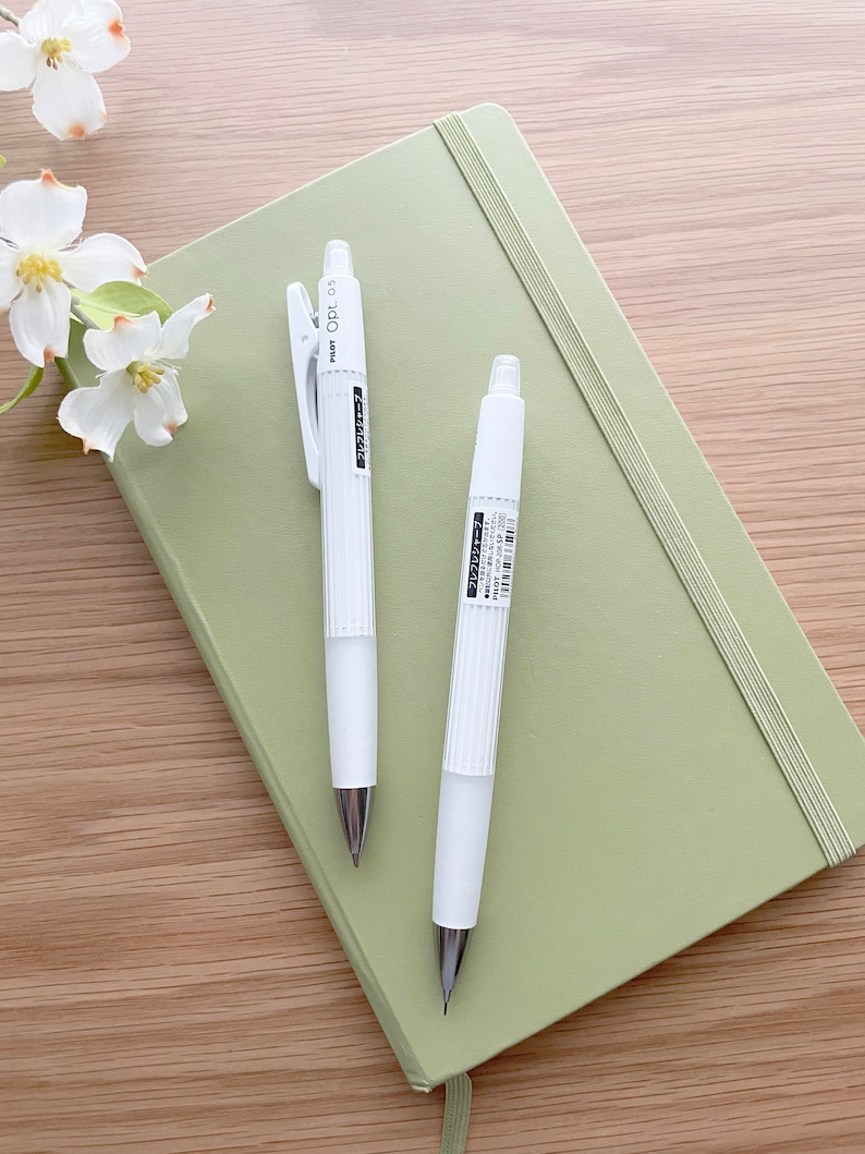 May include: Two white mechanical pencils with a green notebook. The pencils have a black clip and a black inscription that reads "Pilot Opt. 0.5".
