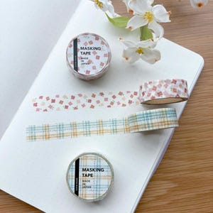 May include: Masking tape rolls with floral and plaid patterns are displayed on an open notebook. One roll has a floral design, and another has a plaid design. The text "MASKING TAPE MADE IN JAPAN" is printed on the rolls.