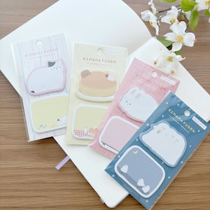 May include: Four packs of "KIMAMA FUSEN" sticky notes with cute animal and food designs. The packs include a cat, a cake, a bunny, and a cloud with a moon and stars. The notes are on a white notebook on a wooden surface.