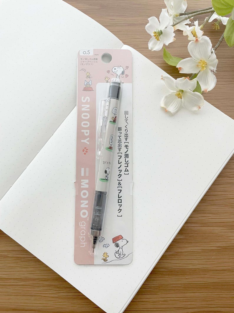 以下が含まれることがあります： スヌーピーのキャラクターデザインが施されたピンクと白のパッケージ。パッケージには、0.5mmの芯の入ったシャープペンシルが入っています。ペンシルは、白いスヌーピーのデザインが施された黒いバレルです。パッケージのテキストには、「Snoopy Mono Graph」と「0.5 mm」と記載されています。