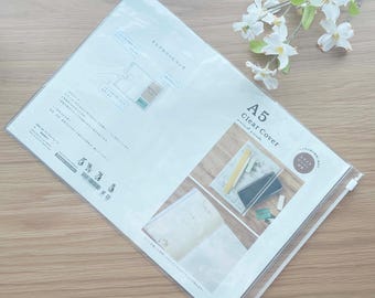 May include: A clear A5 Clear Cover package, sealed with a zipper. The package contains a notebook and stationery. The text "A5 Clear Cover" is printed on the package. A sprig of white flowers rests on the wooden surface.