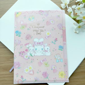 May include: A pink file folder featuring a repeating pattern of flowers, bows, and cartoon bunnies. The folder has the text "Cinnamoroll Miki Taka" and is resting on a white notebook. A pink ribbon is attached to the folder.