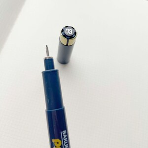 May include: A blue and gold Sakura Pigma Micron pen with a black cap and a silver tip. The pen has a number "03" printed on the cap.