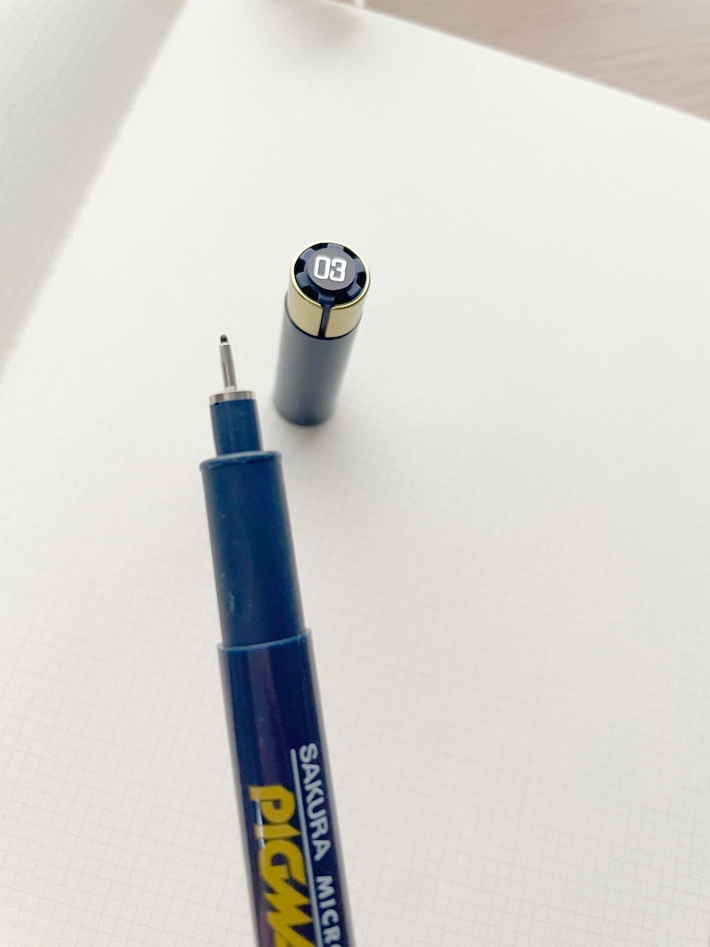 May include: A blue and gold Sakura Pigma Micron pen with a black cap and a silver tip. The pen has a number "03" printed on the cap.