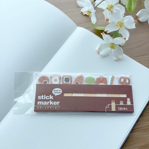 May include: A package of stick markers with food-themed designs, including toast, eggs, and pastries. The package is brown with white text and the words "stick marker" are visible. The markers are on a white dotted notebook.