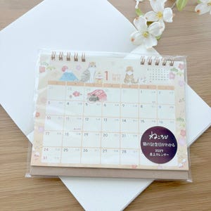 May include: A 2027 desk calendar with a spiral binding, featuring illustrations of cats, Mount Fuji, and floral motifs. The calendar page shows January with a grid of dates and a circular sticker with Japanese text that reads 'ねころび 猫の記念日がわかる 2027 卓上カレンダー'.