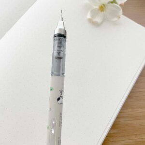 以下が含まれることがあります： 透明なバレルと銀色の先端を持つ白い機械式鉛筆。鉛筆は緑と白のデザインで、漫画の犬が描かれています。鉛筆の側面には「Snoopy」という文字が印刷されています。