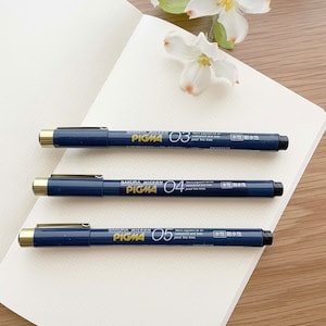 May include: Three black and blue Sakura Micron Pigma pens with gold accents. The pens are labeled 03, 04, and 05. The text "水性耐水性" is printed on each pen, which translates to "water-resistant".
