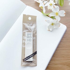 May include: A close-up of a white Uni mechanical pencil with a 0.5 HB lead. The pencil is packaged in a clear plastic bag with a brown paper label that says "Uni 0.5 HB" and "Limited edition".