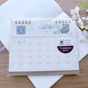 May include: A 2027 desk calendar for January featuring a red panda illustration, a spiral binding, and a circular sticker that reads Ressa Panda Books Calendar 2027. The calendar grid displays dates and small snowflake and snowman graphics.