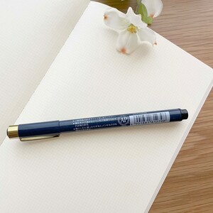 May include: A blank, white, gridded notebook with a blue pen on top. The pen has gold accents and a black tip. The pen has Japanese text on it, including the words "AP" and "4 901881 812798".