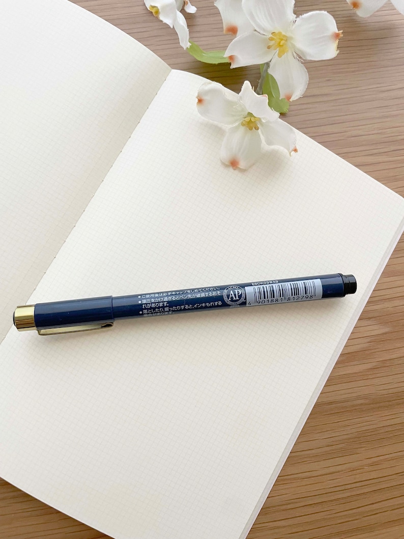 May include: A blank, white, gridded notebook with a blue pen on top. The pen has gold accents and a black tip. The pen has Japanese text on it, including the words "AP" and "4 901881 812798".