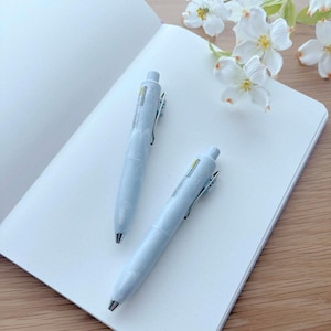 May include: Two pale blue pens with silver detailing lie on an open, dot-grid notebook. The notebook is white, and the background includes white flowers and a wooden surface. The pens are a light blue colour.