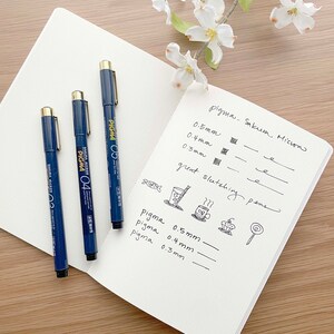 May include: Three blue Sakura Pigma Micron pens with gold accents and black ink. The pens are labeled 0.3mm, 0.4mm, and 0.5mm. The pens are on a white notebook with handwritten notes about the pens and their uses.