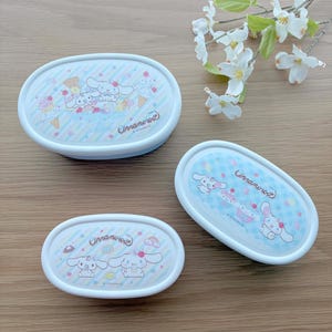 May include: Three oval-shaped, white and blue food containers. Each container lid features a cartoon illustration of a white dog-like character with desserts and the text "Cinnamoroll". The containers are on a light brown surface.