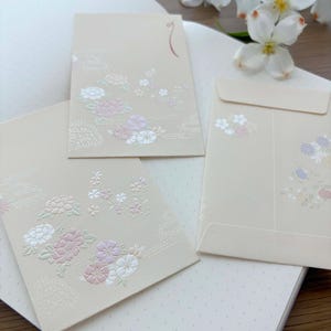May include: Beige stationery set featuring floral designs in pink, white, and lavender. The set includes envelopes and a notepad with a dotted pattern. A sprig of white flowers is in the background.