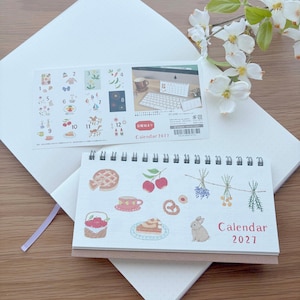 May include: A 2027 desk calendar with a spiral binding, featuring watercolor-style illustrations of a pie, cherries, herbs, a basket of apples, a cake, and a rabbit. It includes a separate instruction sheet with numbered icons and product details.