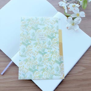 May include: A white notebook with a floral patterned cover featuring yellow and green mimosa branches. The cover has the text "from the Garden of Mimosa". A yellow tabbed divider is visible. A sprig of white flowers is in the background.