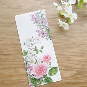 May include: A white card with a floral design featuring pink and white roses, green leaves, and white flowers. The card is on a light brown wooden surface.