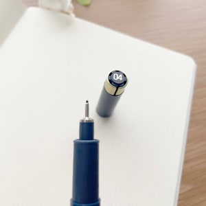 May include: A blue and silver fineliner pen with a black cap and the number "04" printed on the cap. The pen is lying on a white notebook.